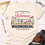 Women's funny woodworking graphic tee shirt for female woodworkers, makers, and DIY builders — Sawdust & Ponytails. Cream t-shirt with "Never Underestimate A Woman Woodworking In Her Workshop" text and tools graphic.
