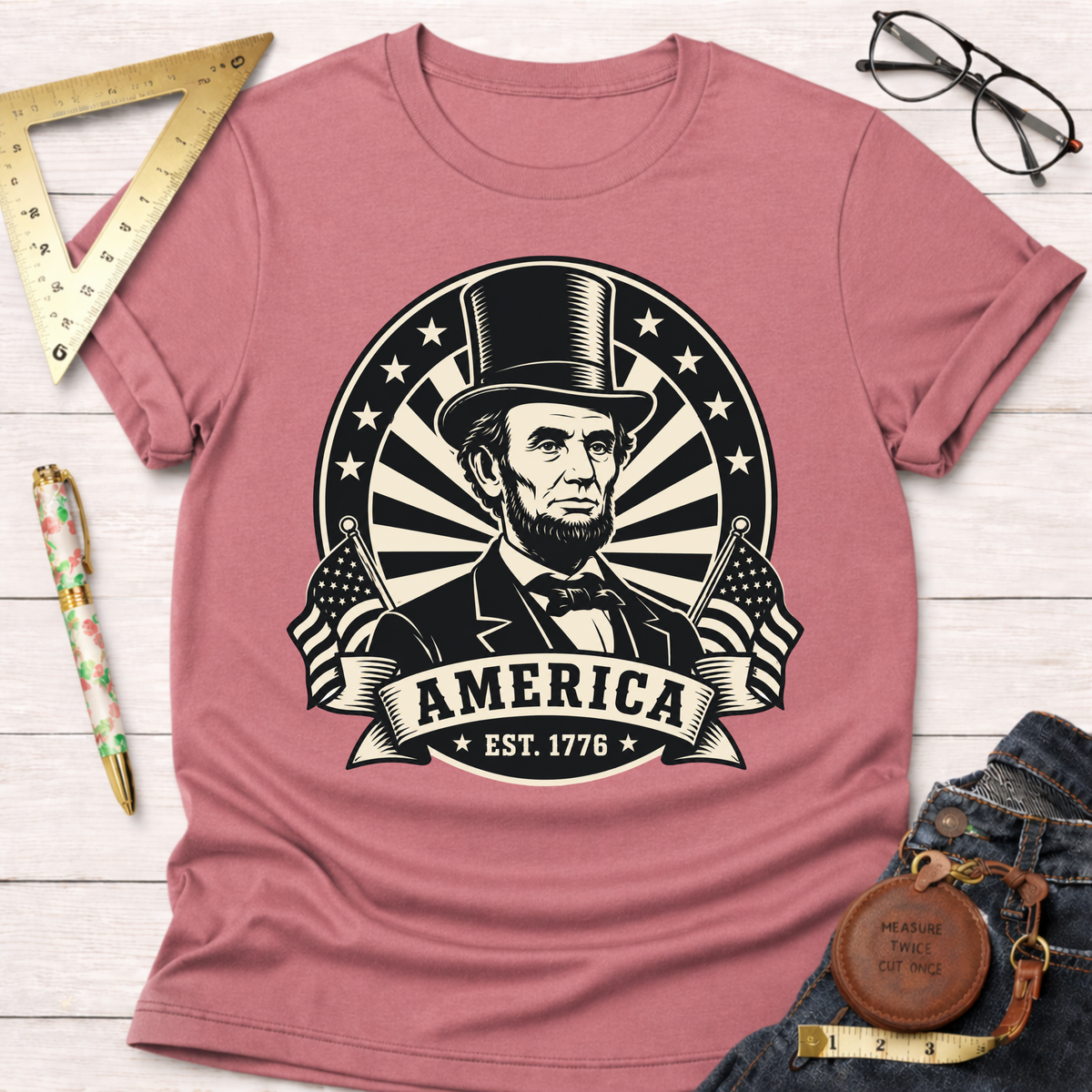 Pink cotton t-shirt with a black graphic of Abraham Lincoln, "AMERICA EST. 1776," and American flags.