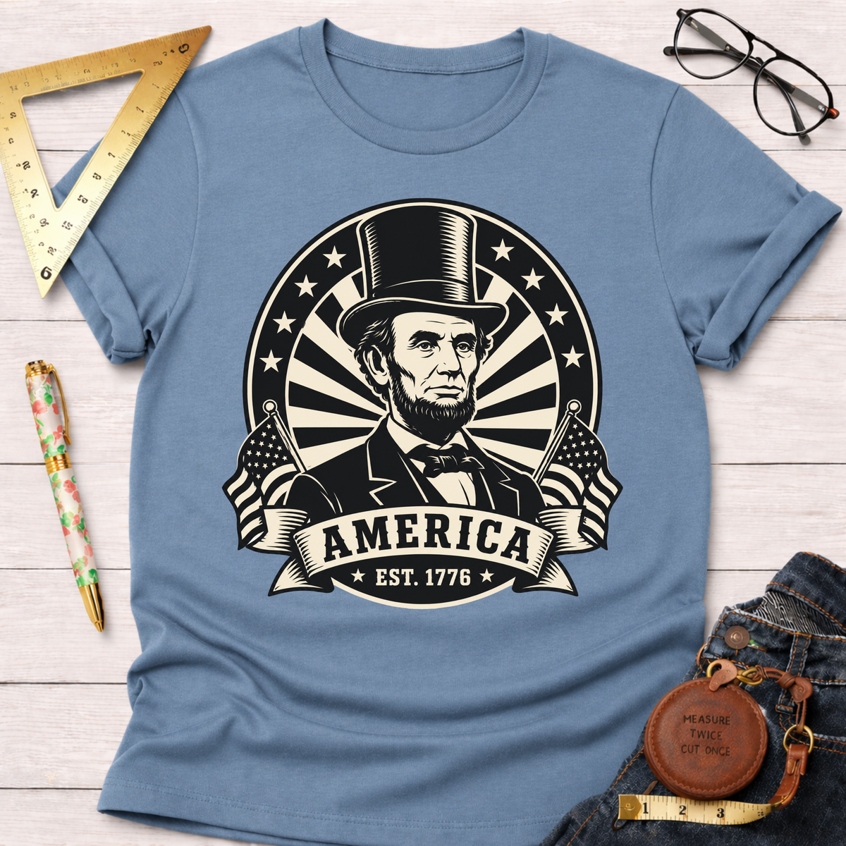 Light blue cotton t-shirt featuring a black and white Abraham Lincoln design with "AMERICA EST. 1776" and flags.