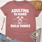 Women's funny woodworking graphic tee shirt for female woodworkers, makers, and DIY builders — Sawdust & Ponytails. Mauve cotton t-shirt with "Adulting Is Hard So I Build Things" text and crossed hammers.