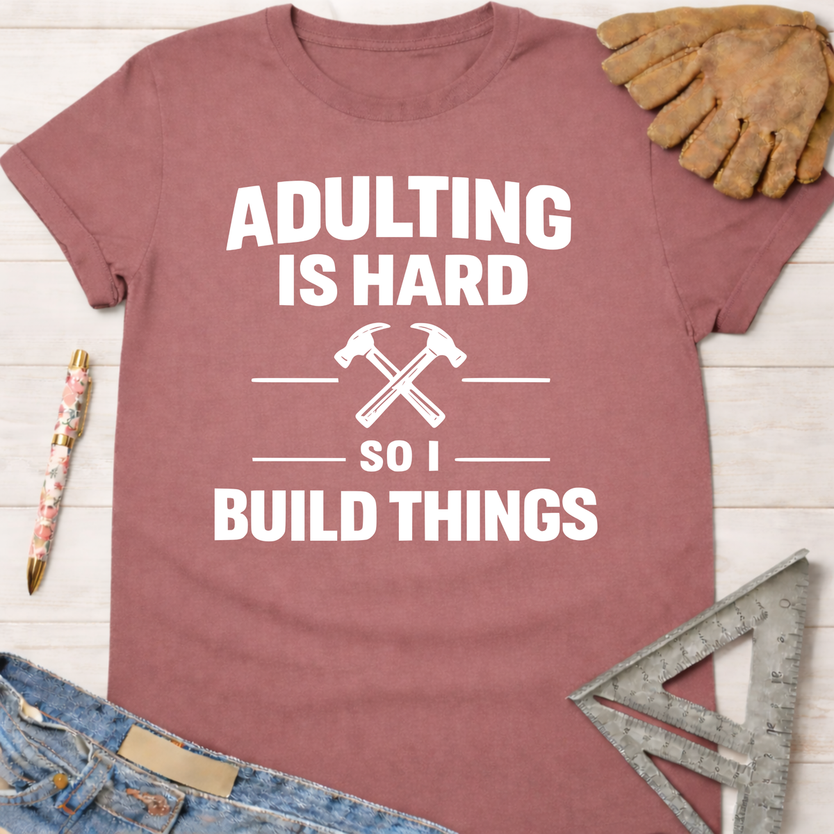Women's funny woodworking graphic tee shirt for female woodworkers, makers, and DIY builders — Sawdust & Ponytails. Mauve cotton t-shirt with "Adulting Is Hard So I Build Things" text and crossed hammers.