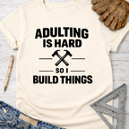 Women's funny woodworking graphic tee shirt for female woodworkers, makers, and DIY builders — Sawdust & Ponytails. Cream t-shirt with "Adulting Is Hard So I Build Things" text and crossed hammers graphic.