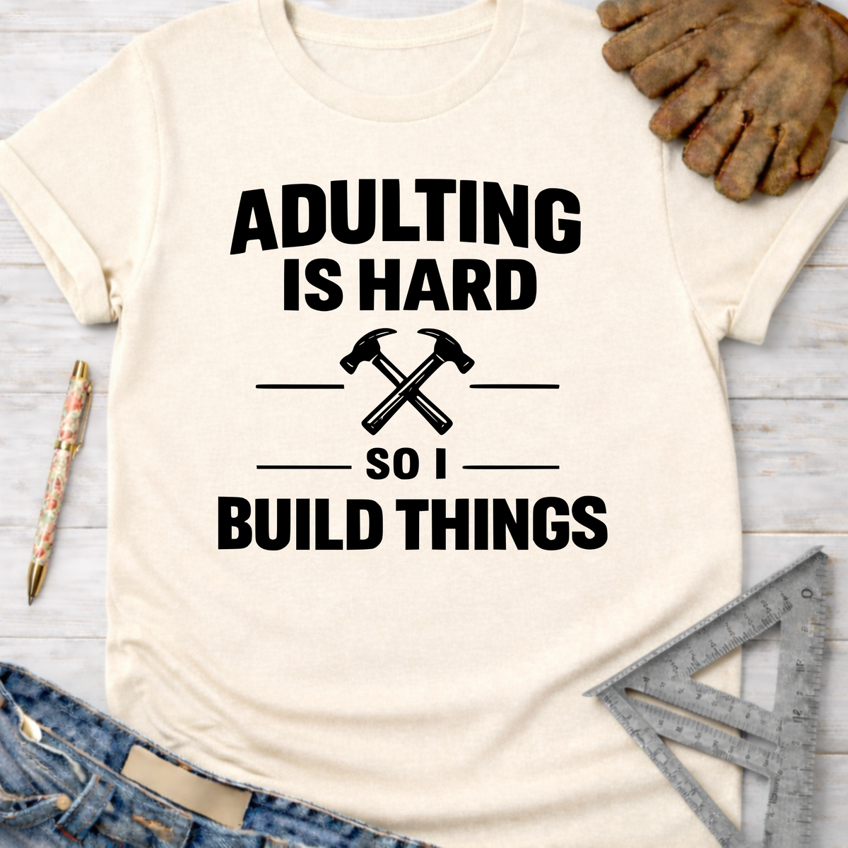 Women's funny woodworking graphic tee shirt for female woodworkers, makers, and DIY builders — Sawdust & Ponytails. Cream t-shirt with "Adulting Is Hard So I Build Things" text and crossed hammers graphic.