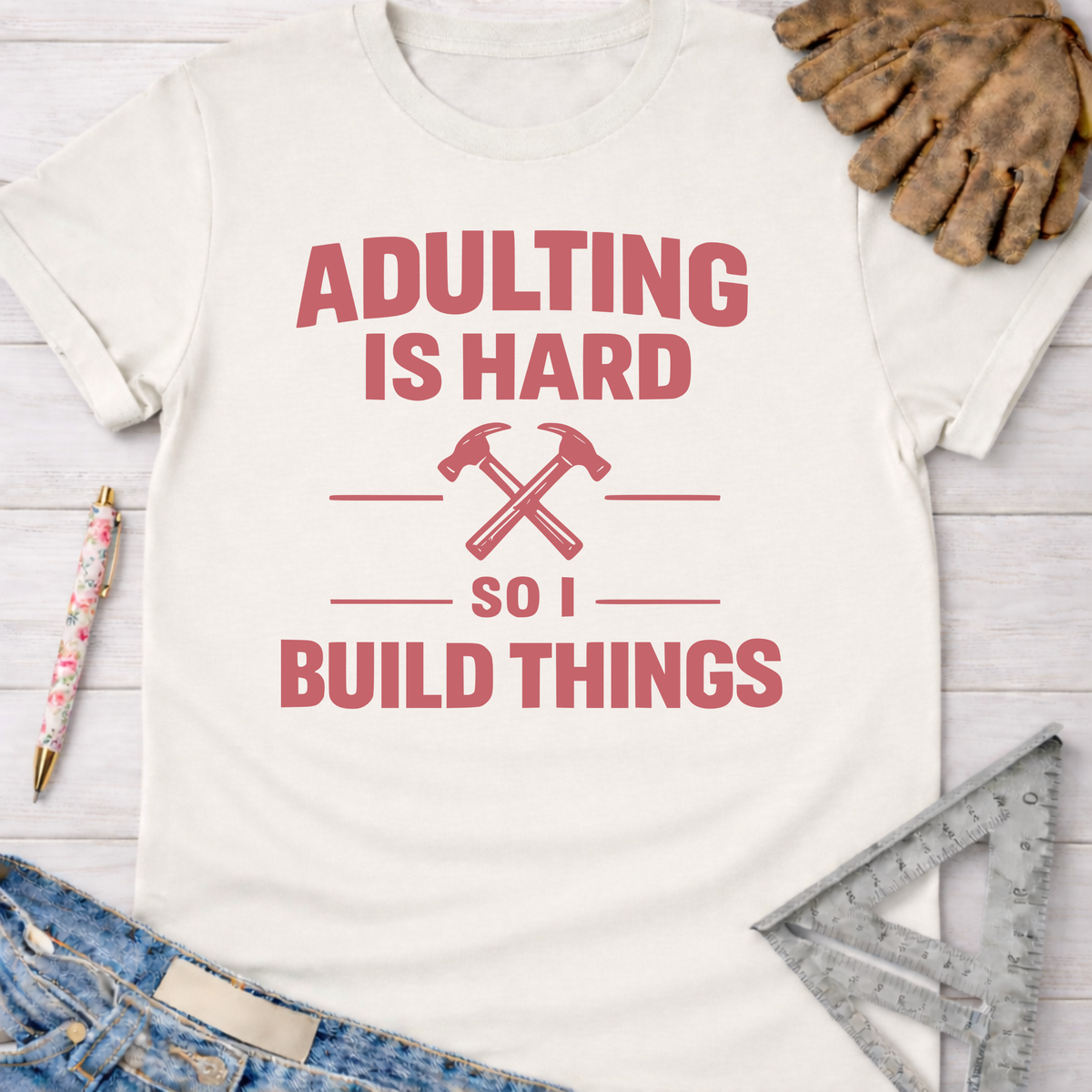 Women's funny woodworking graphic tee shirt for female woodworkers, makers, and DIY builders — Sawdust & Ponytails. Cream t-shirt with "Adulting Is Hard So I Build Things" in pink, featuring crossed hammers.