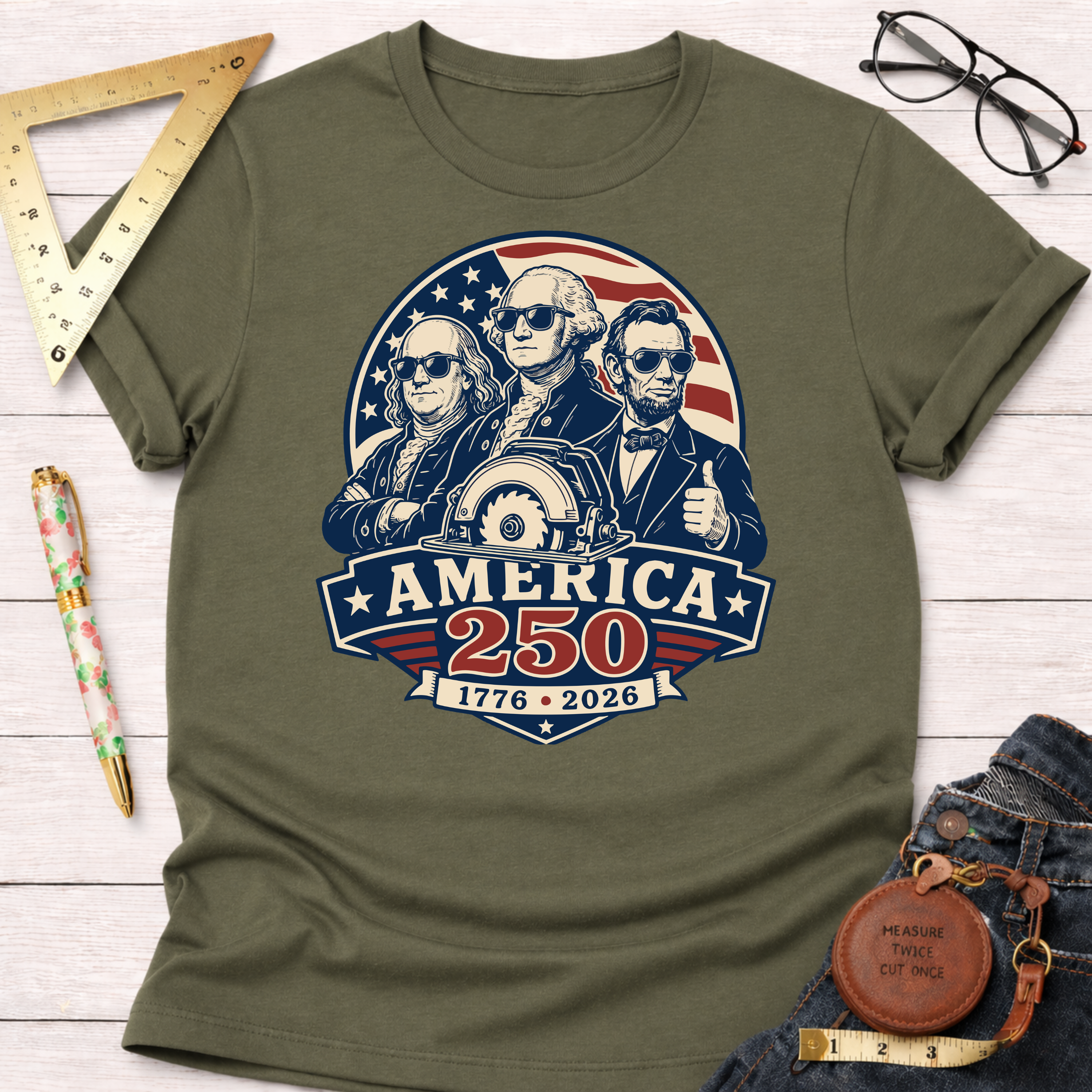 Olive green cotton t-shirt with a graphic of Benjamin Franklin, George Washington, and Abraham Lincoln in sunglasses, with a saw blade, "America 250," and "1776-2026.