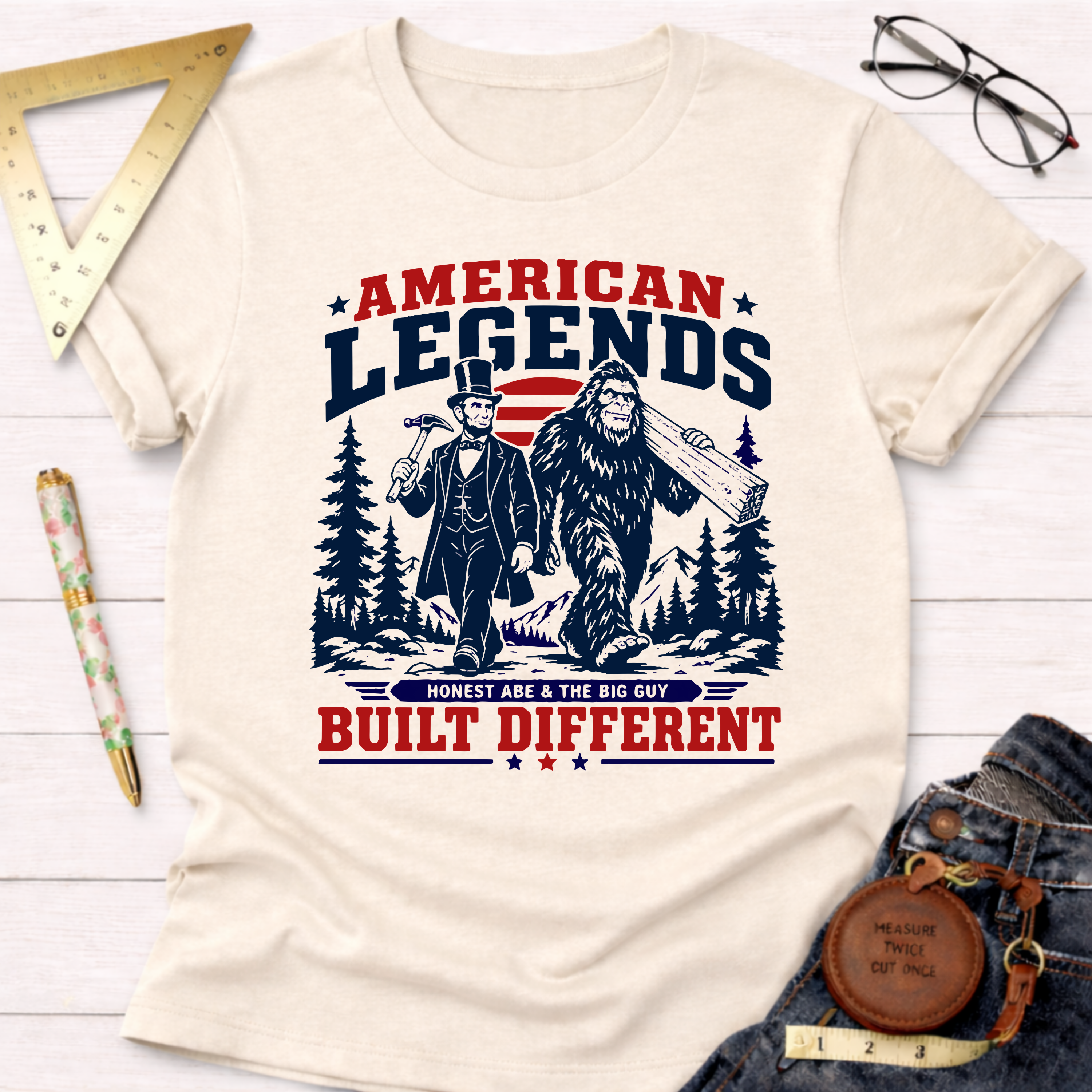 Cream cotton t-shirt with "American Legends Built Different" graphic featuring Abraham Lincoln and Bigfoot carrying wood.