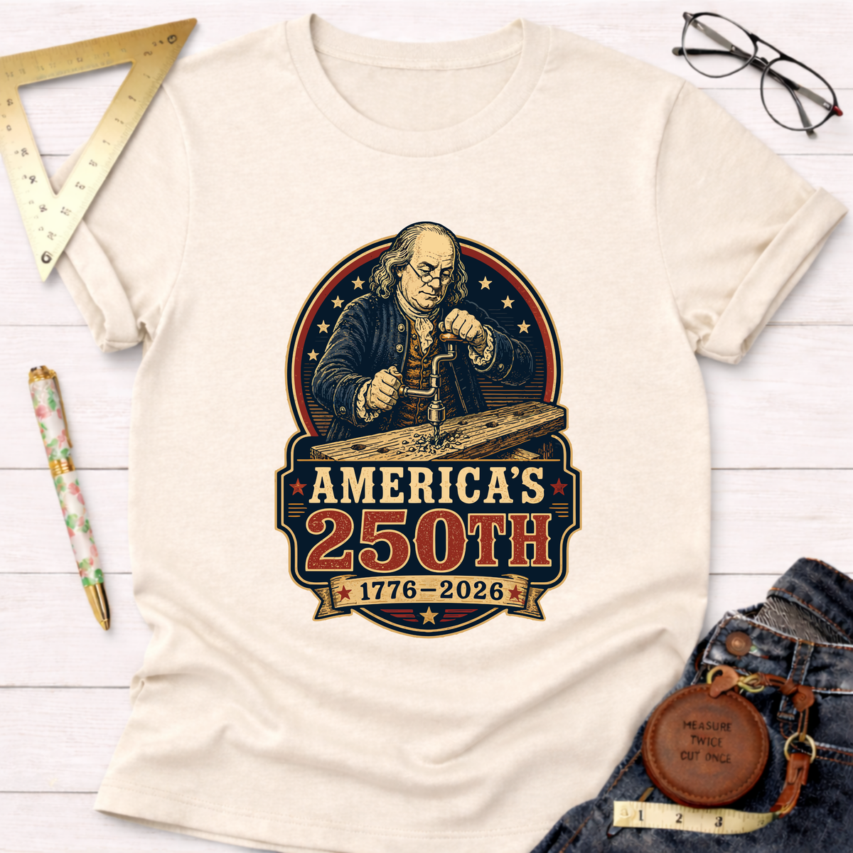 Cream cotton t-shirt with a graphic of Benjamin Franklin woodworking, "America's 250th 1776-2026" text.
