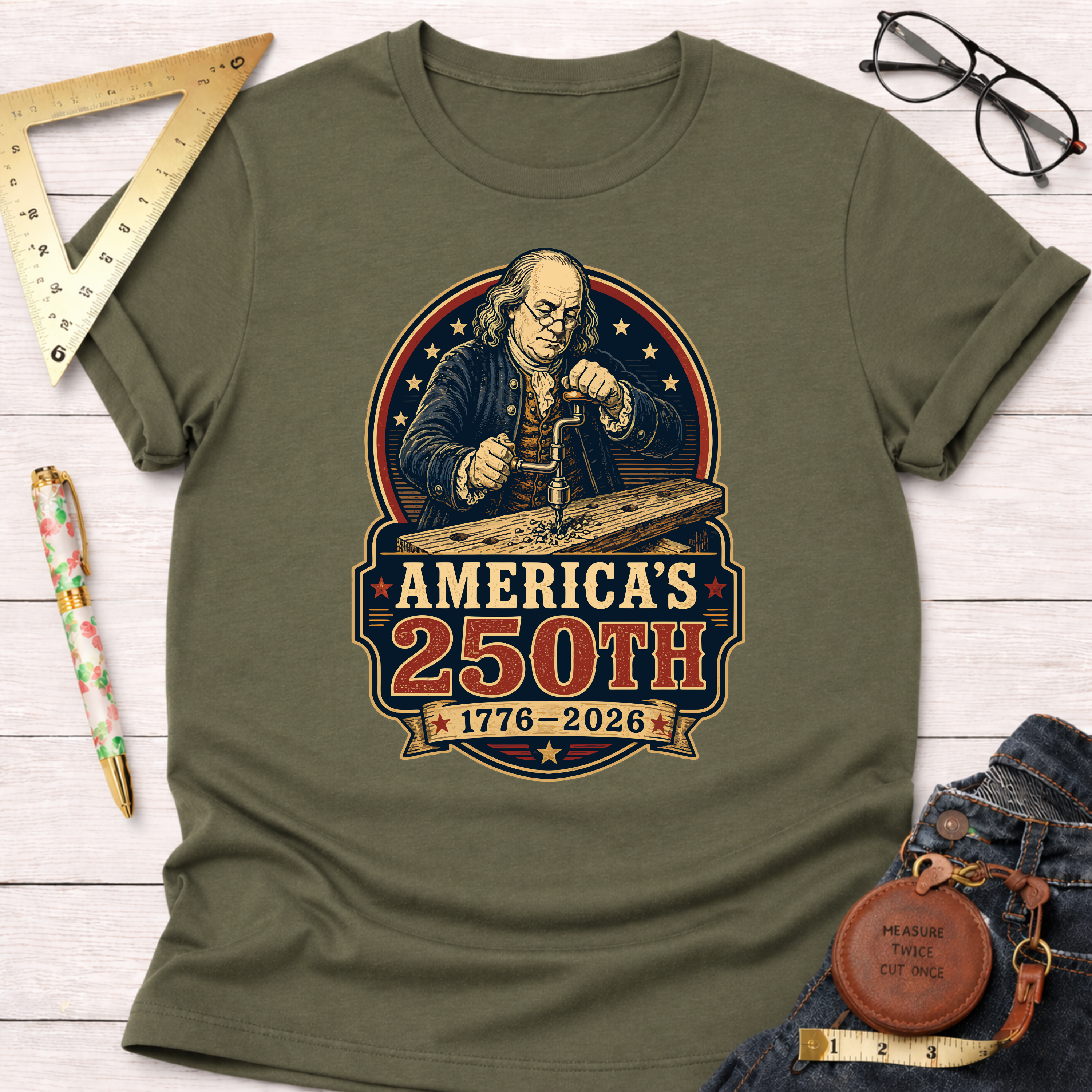Olive green cotton t-shirt with a graphic of Benjamin Franklin drilling wood, celebrating America's 250th.