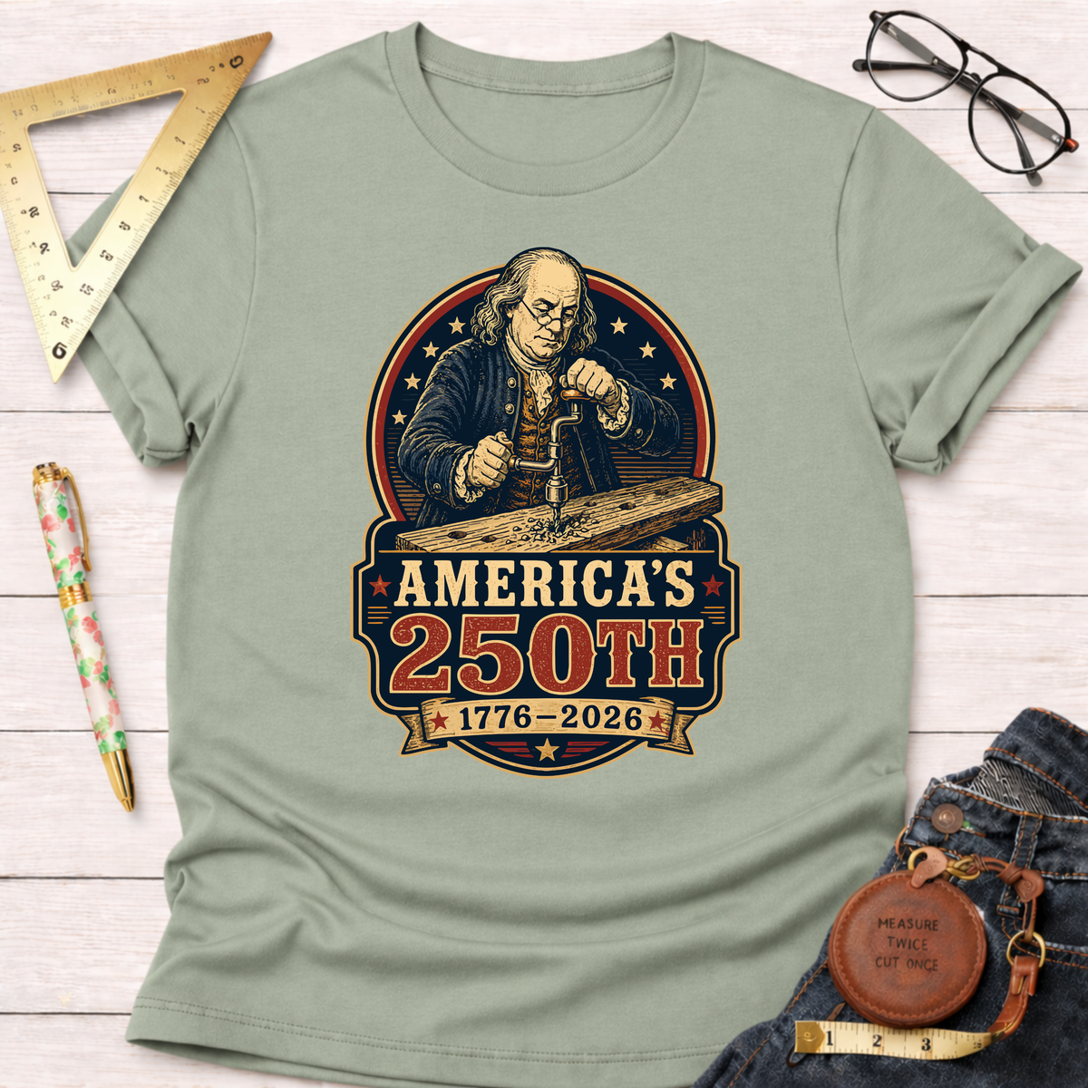 Sage green cotton t-shirt with a graphic of Benjamin Franklin woodworking, "America's 250th 1776-2026".