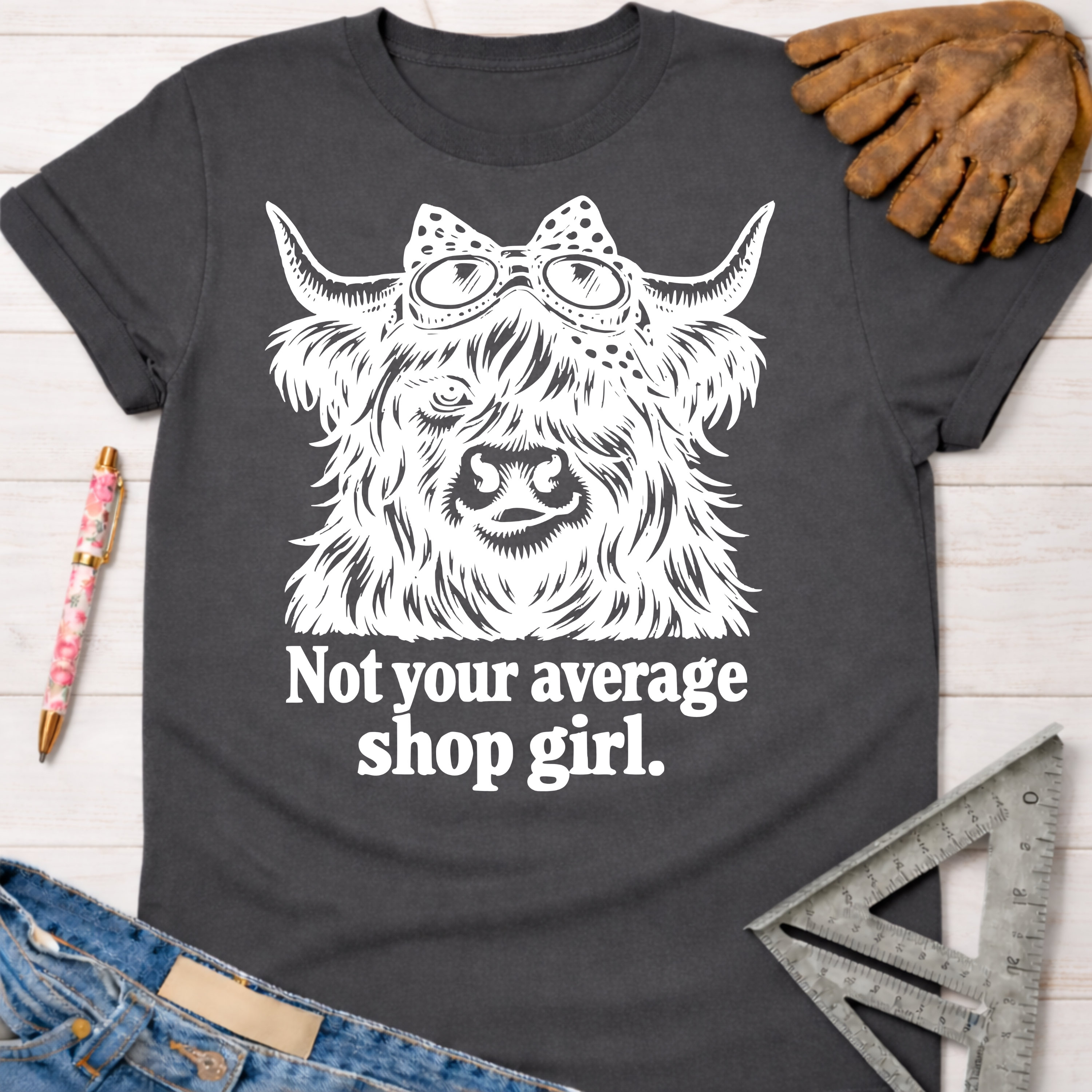 Women's funny woodworking graphic tee shirt for female woodworkers, makers, and DIY builders — Sawdust & Ponytails. Dark gray crewneck t-shirt with a white graphic of a Highland cow wearing a bow and goggles, above "Not your average shop girl.