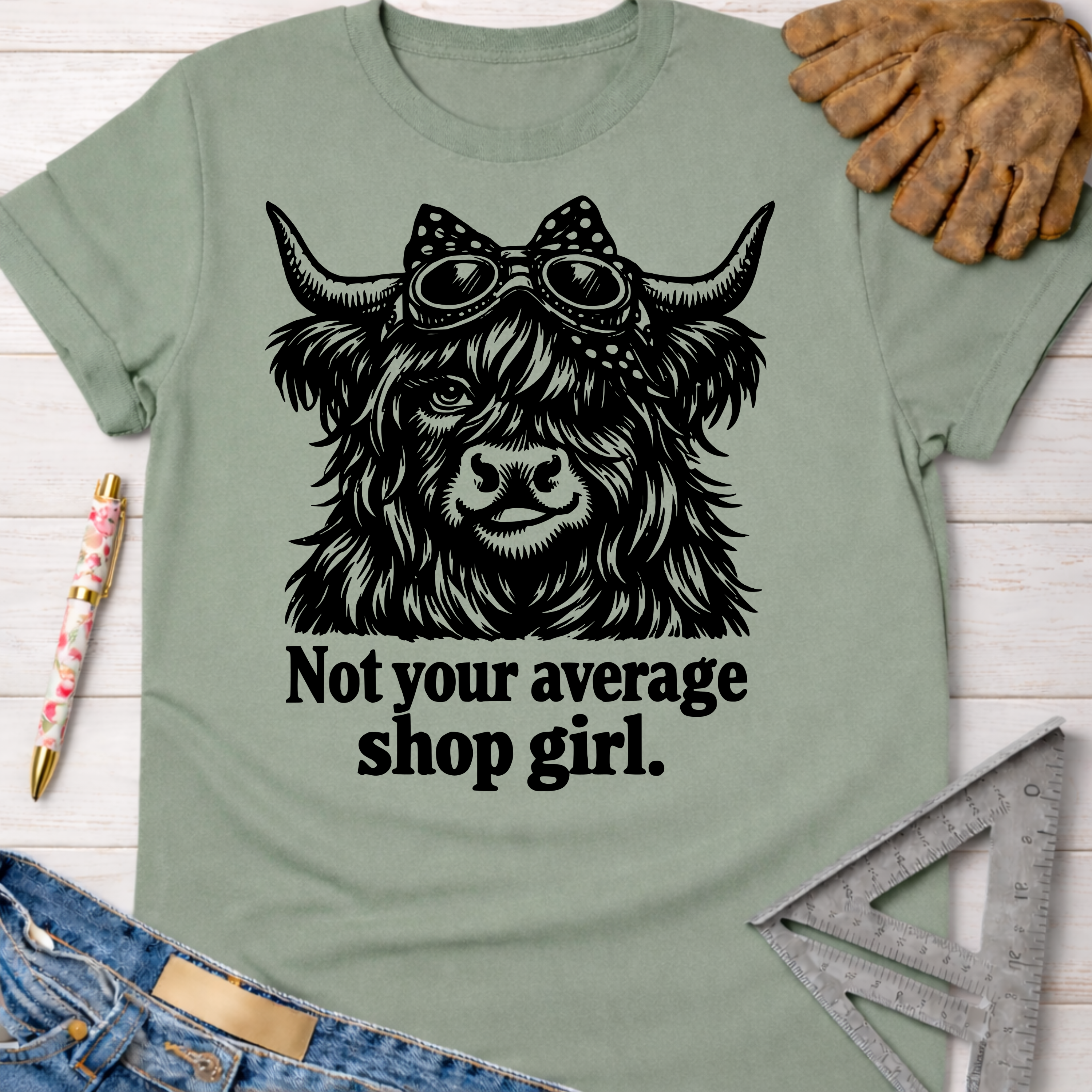Women's funny woodworking graphic tee shirt for female woodworkers, makers, and DIY builders — Sawdust & Ponytails. Sage green cotton t-shirt with a black graphic of a Highland cow wearing goggles and a polka dot bow, above the text "Not your average shop girl.
