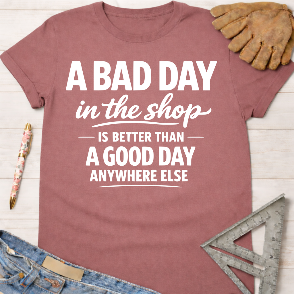 Women's funny woodworking graphic tee shirt for female woodworkers, makers, and DIY builders — Sawdust & Ponytails. Mauve cotton tee with "A BAD DAY in the shop IS BETTER THAN A GOOD DAY ANYWHERE ELSE" in white text.