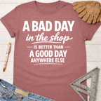 Women's funny woodworking graphic tee shirt for female woodworkers, makers, and DIY builders — Sawdust & Ponytails. Mauve cotton tee with "A BAD DAY in the shop IS BETTER THAN A GOOD DAY ANYWHERE ELSE" in white text.