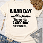 Women's funny woodworking graphic tee shirt for female woodworkers, makers, and DIY builders — Sawdust & Ponytails. Cream t-shirt with "A BAD DAY in the shop IS BETTER THAN A GOOD DAY ANYWHERE ELSE" in black text.