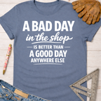 Women's funny woodworking graphic tee shirt for female woodworkers, makers, and DIY builders — Sawdust & Ponytails. Blue cotton tee with "A BAD DAY in the shop IS BETTER THAN A GOOD DAY ANYWHERE ELSE" text.