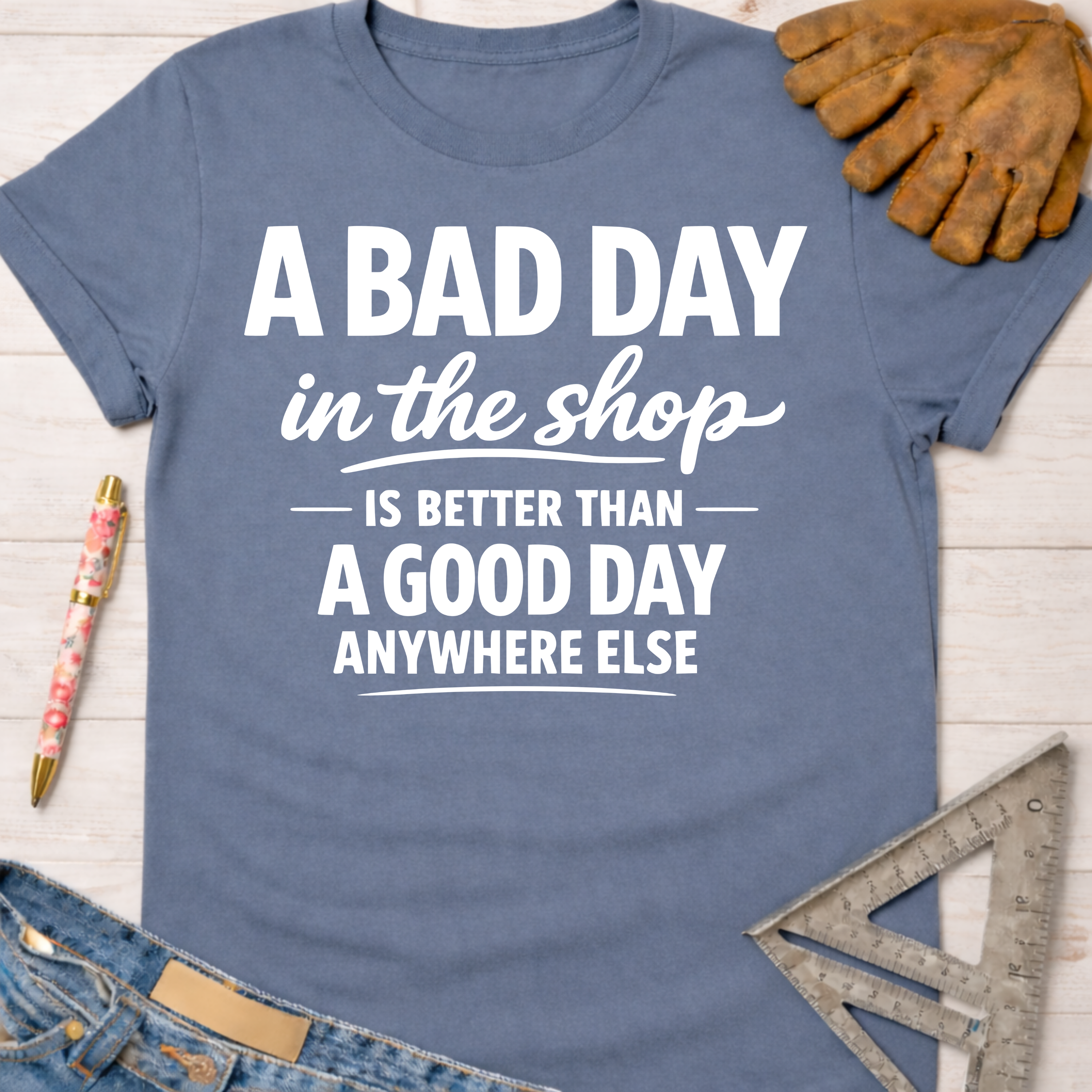 Women's funny woodworking graphic tee shirt for female woodworkers, makers, and DIY builders — Sawdust & Ponytails. Blue cotton tee with "A BAD DAY in the shop IS BETTER THAN A GOOD DAY ANYWHERE ELSE" text.