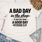 Women's funny woodworking graphic tee shirt for female woodworkers, makers, and DIY builders — Sawdust & Ponytails. White cotton tee with "A BAD DAY in the shop IS BETTER THAN A GOOD DAY ANYWHERE ELSE" in black text.