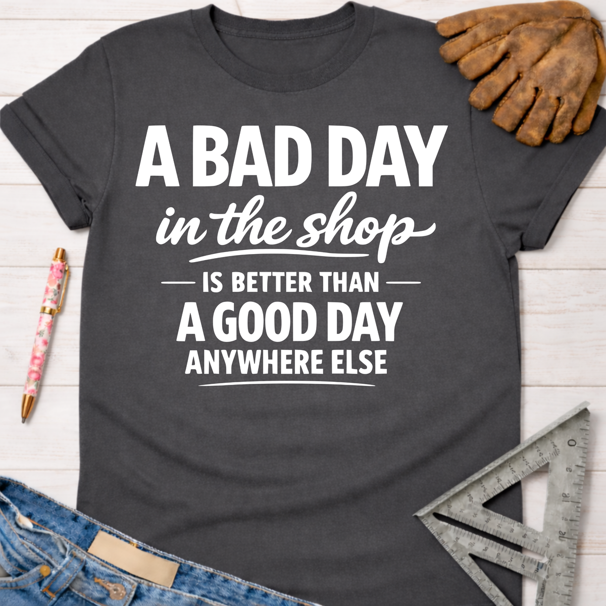 Women's funny woodworking graphic tee shirt for female woodworkers, makers, and DIY builders — Sawdust & Ponytails. Dark gray cotton tee with white text: "A BAD DAY in the shop IS BETTER THAN A GOOD DAY ANYWHERE ELSE.