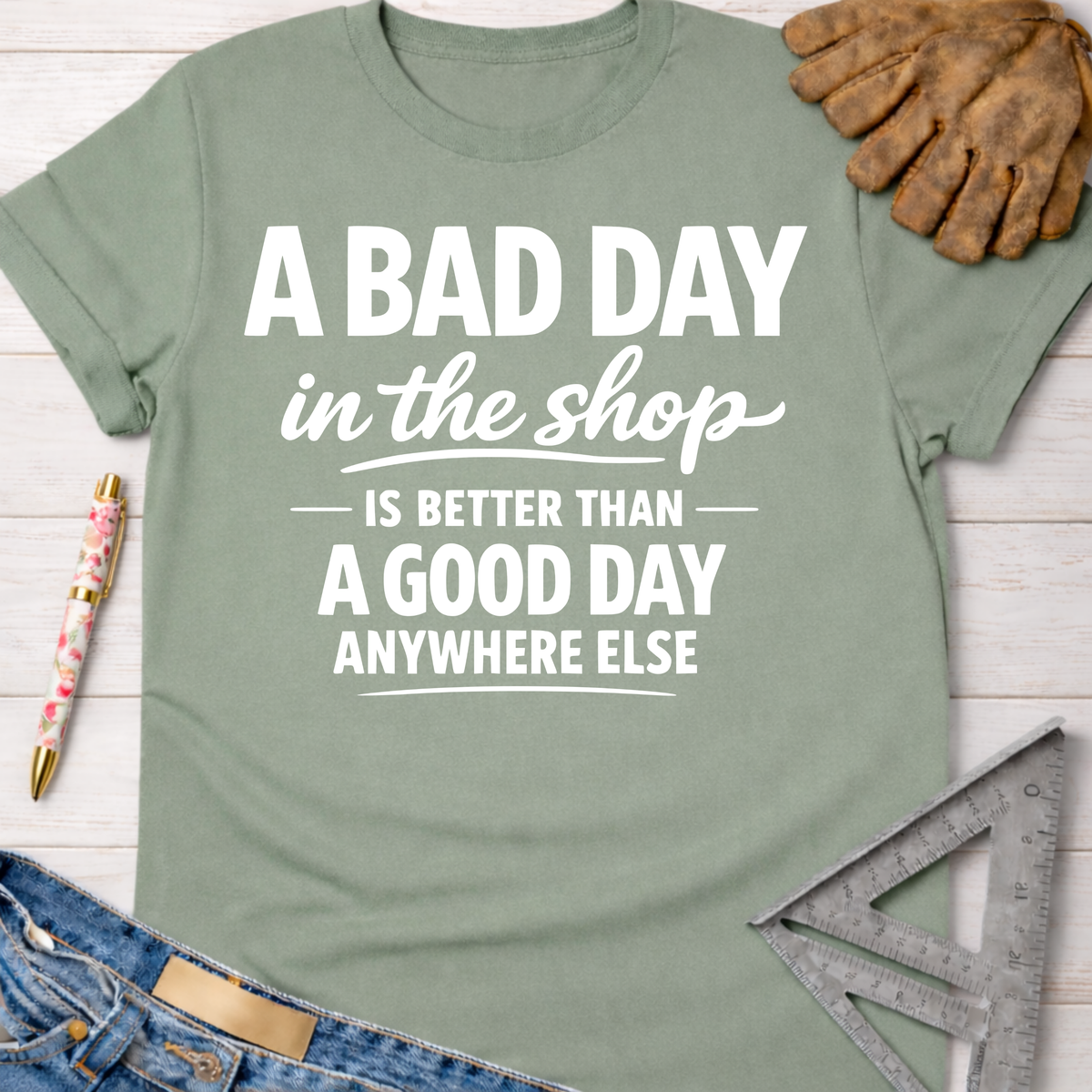 Women's funny woodworking graphic tee shirt for female woodworkers, makers, and DIY builders — Sawdust & Ponytails. Sage green cotton tee with "A BAD DAY in the shop IS BETTER THAN A GOOD DAY ANYWHERE ELSE" text.