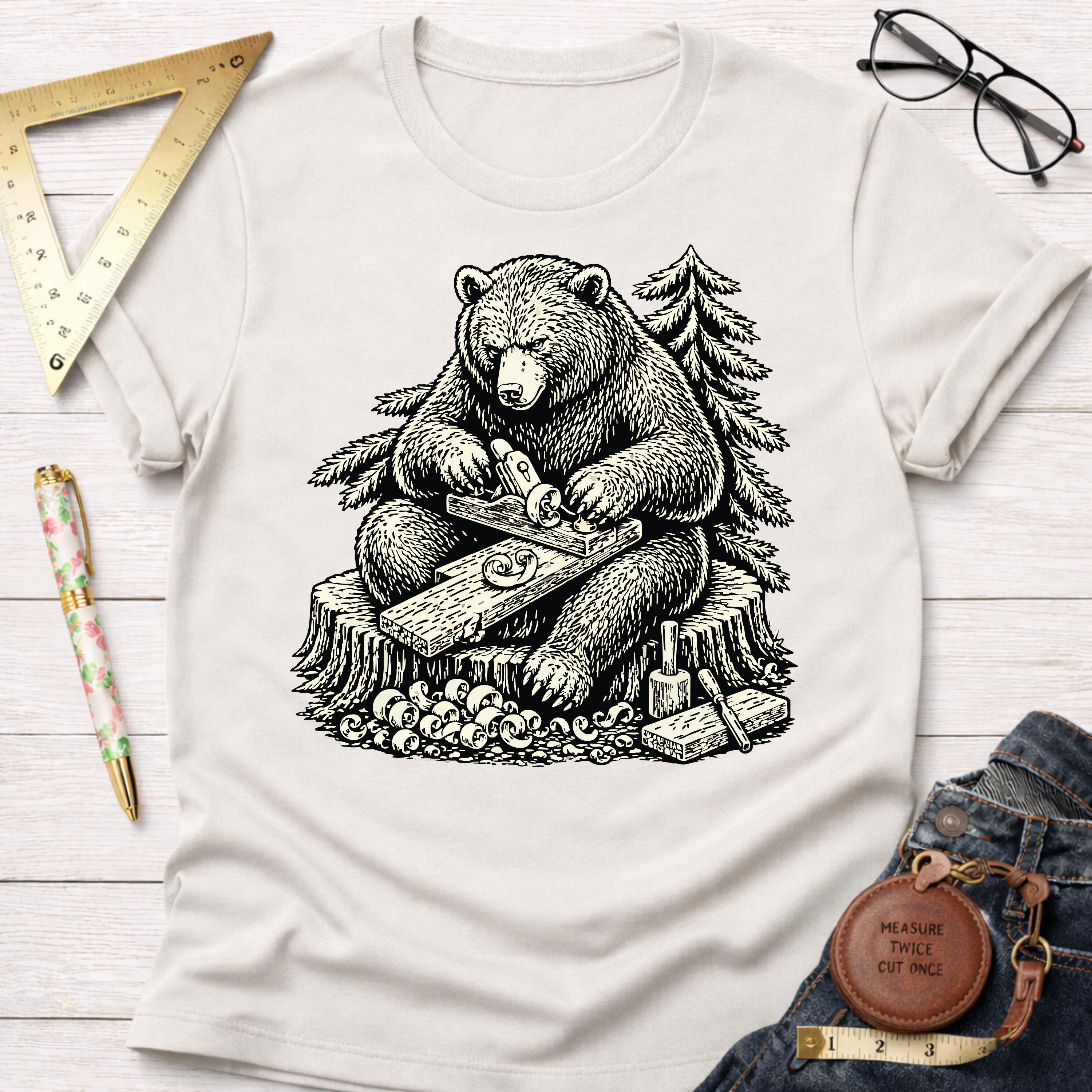 Women's funny woodworking graphic tee shirt for female woodworkers, makers, and DIY builders — Sawdust & Ponytails. Cream t-shirt with a black graphic of a bear planing wood on a tree stump.