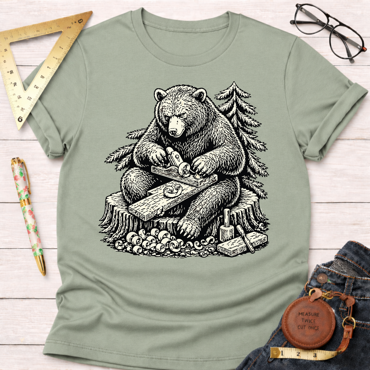 Women's funny woodworking graphic tee shirt for female woodworkers, makers, and DIY builders — Sawdust & Ponytails. Sage green cotton t-shirt with a black graphic of a bear planing wood.