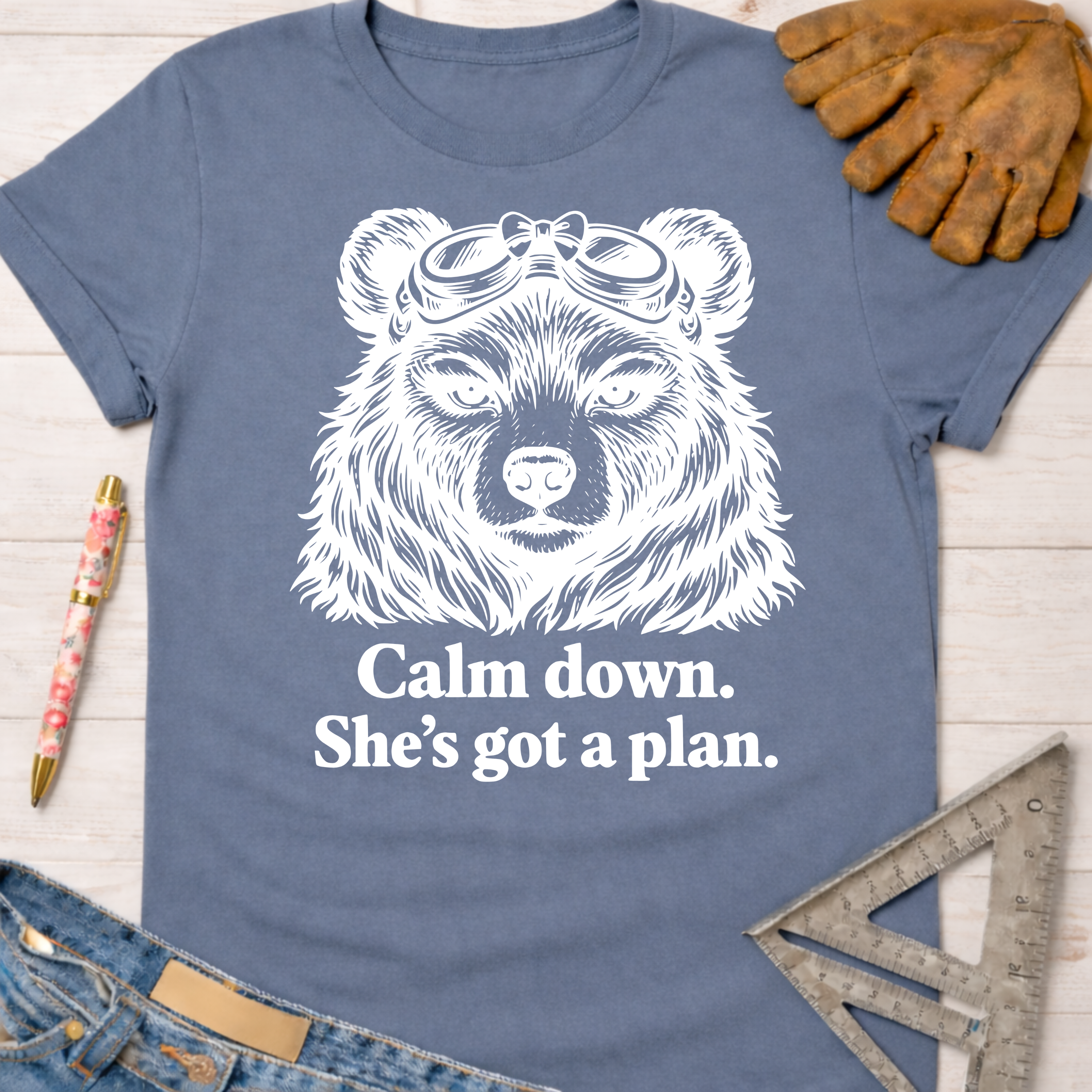 Women's funny woodworking graphic tee shirt for female woodworkers, makers, and DIY builders — Sawdust & Ponytails. Blue cotton tee with white bear in goggles graphic and "Calm down. She's got a plan." text.
