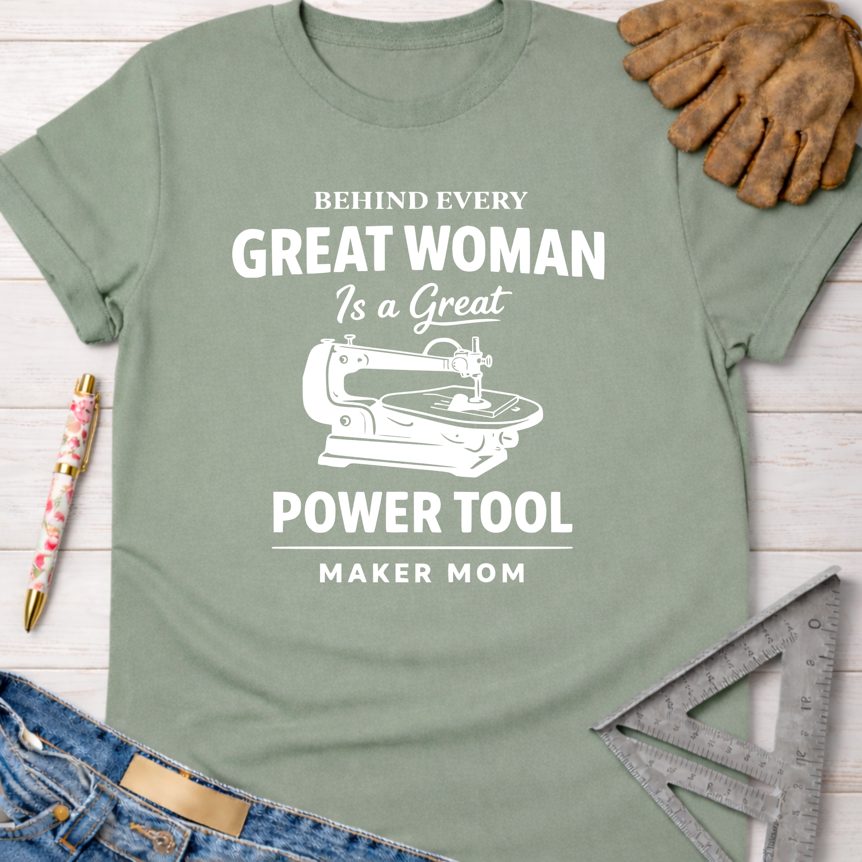 Women's funny woodworking graphic tee shirt for female woodworkers, makers, and DIY builders — Sawdust & Ponytails. Sage green cotton tee with "Behind Every Great Woman Is a Great Power Tool" text and a sewing machine graphic.