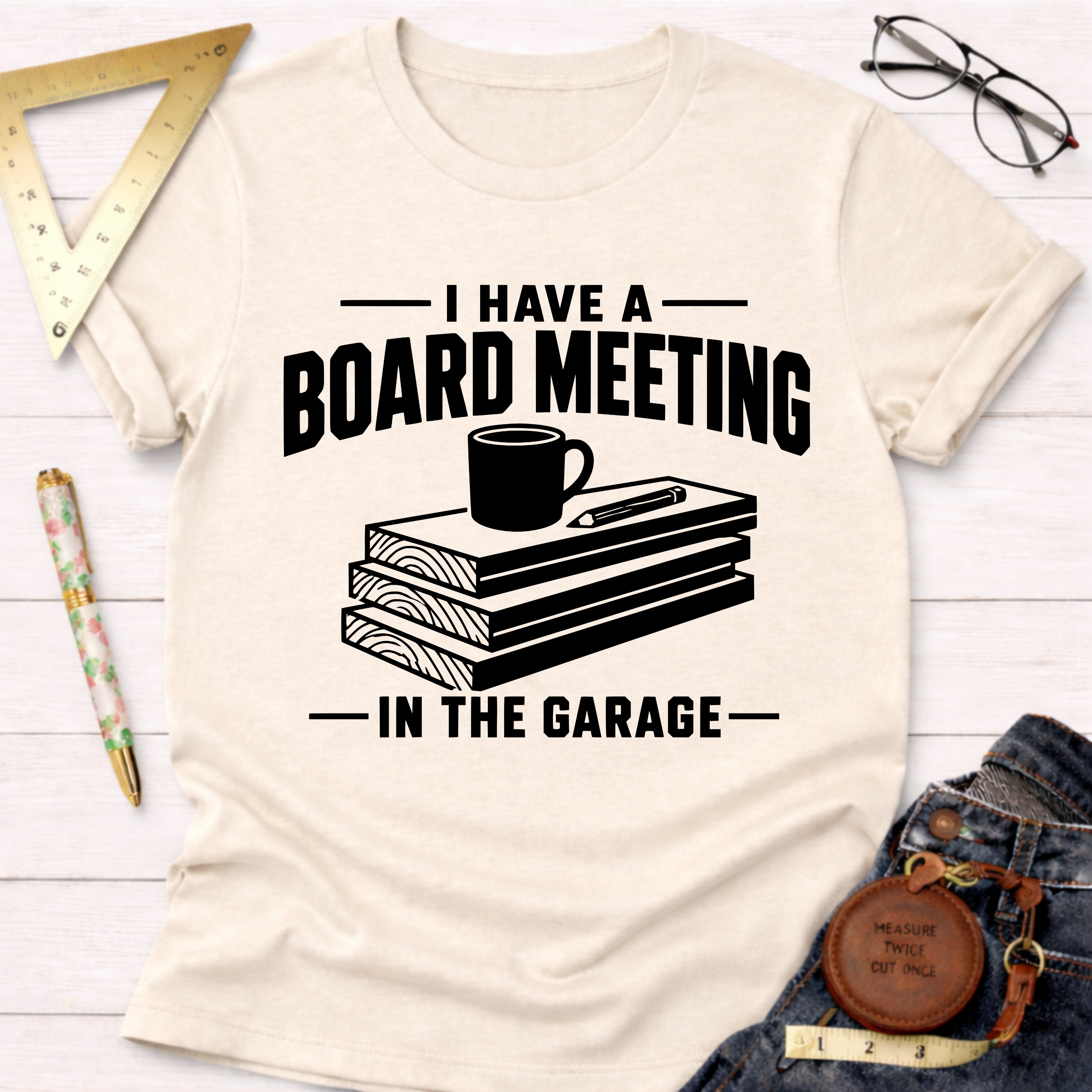 Cream t-shirt with "I Have A Board Meeting In The Garage" text, stacked wood, coffee cup, and pencil graphic.