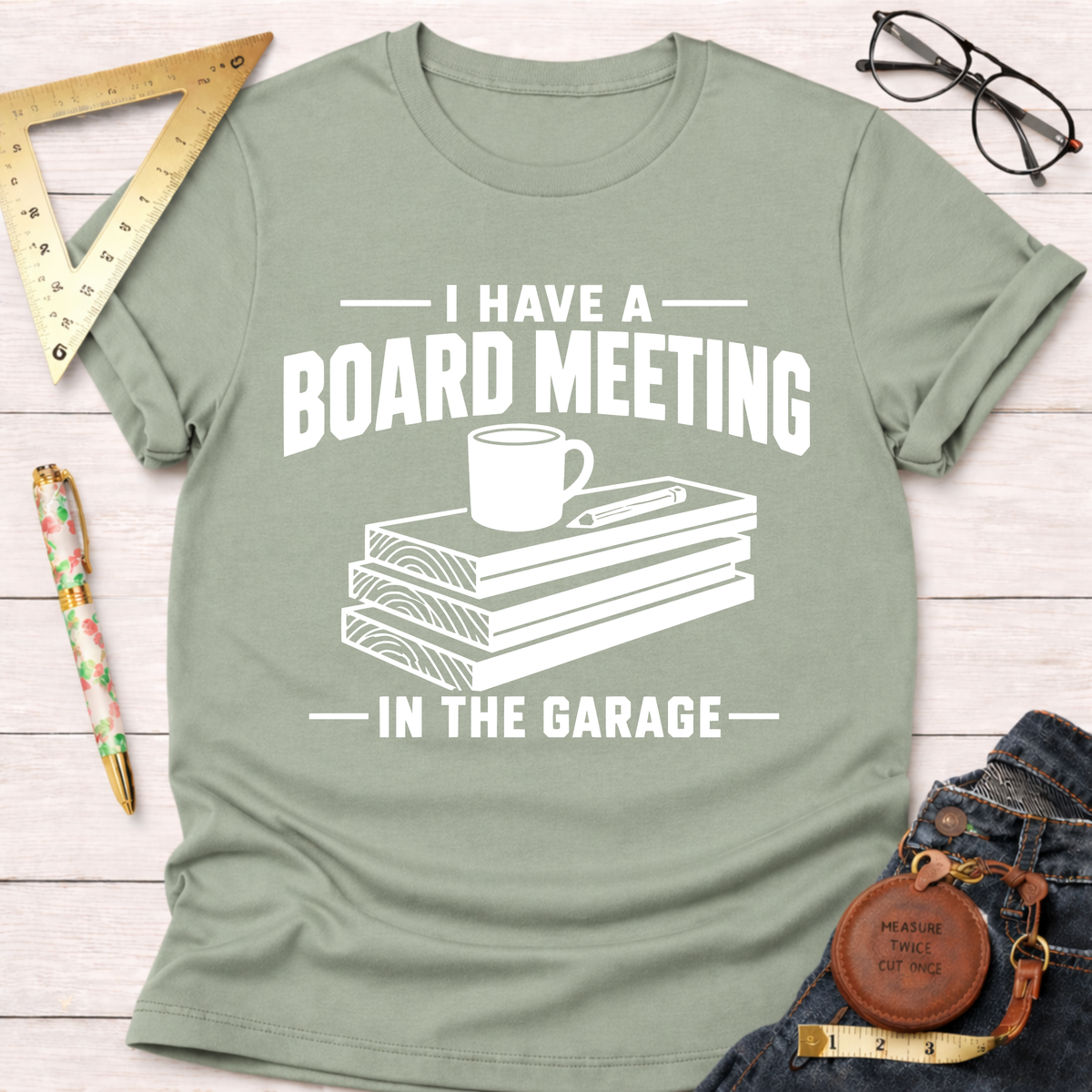Sage green t-shirt with "I Have A Board Meeting In The Garage" text, wood stack, coffee cup, and pencil graphic.