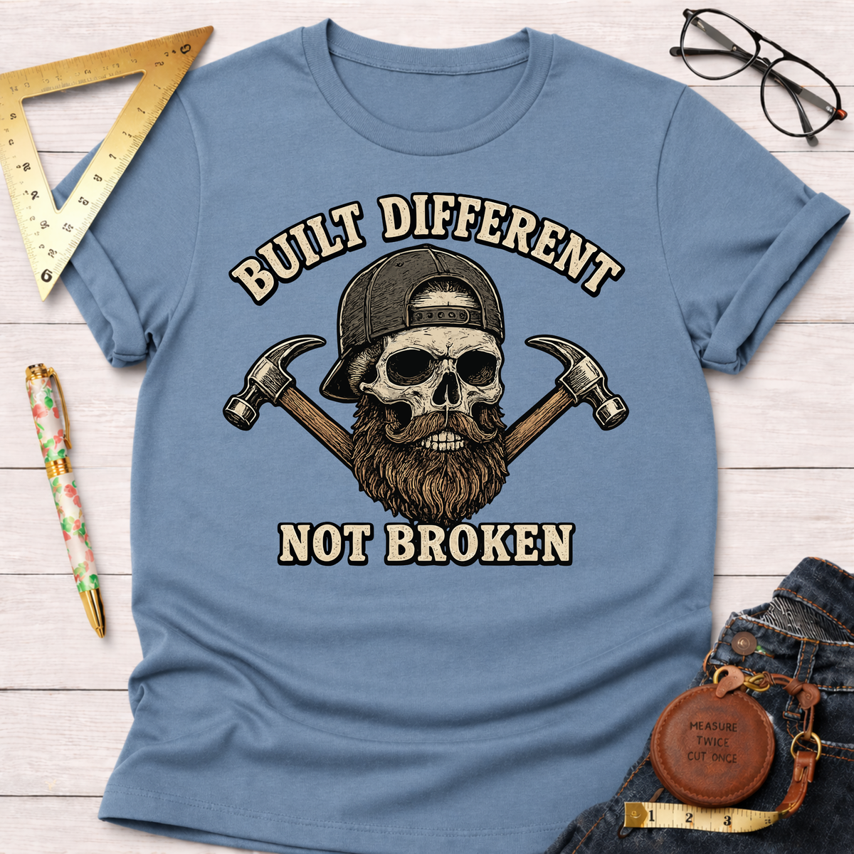 Light blue "Built Different Not Broken" t-shirt with a skull wearing a cap and beard, flanked by hammers.