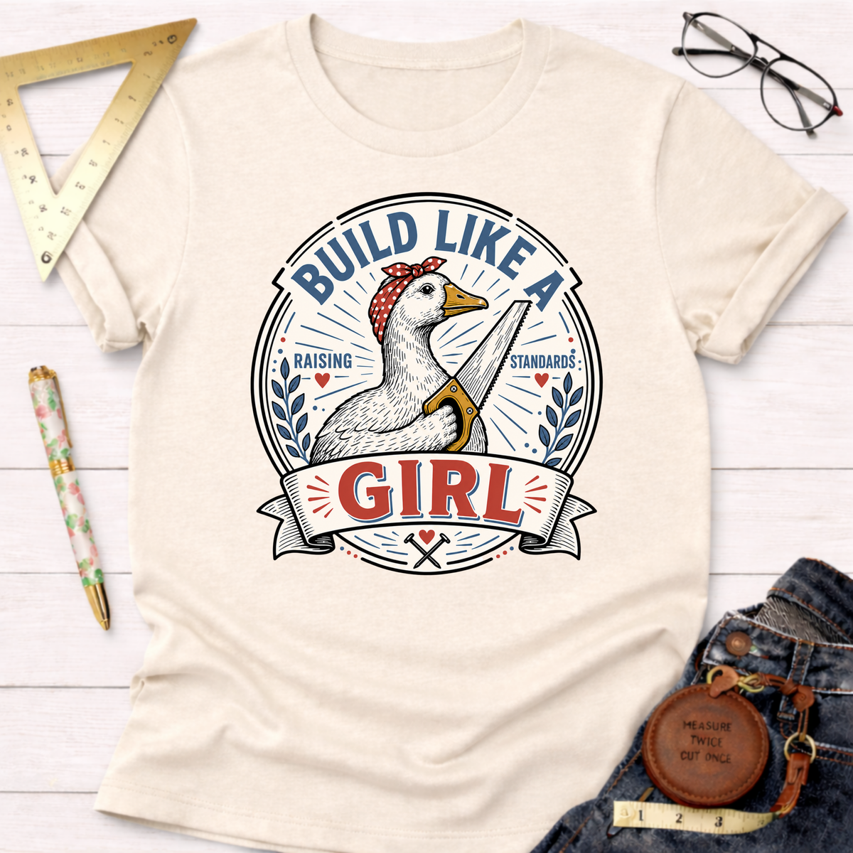 Cream "Build Like A Girl" t-shirt with a goose wearing a bandana and holding a saw, surrounded by tools.