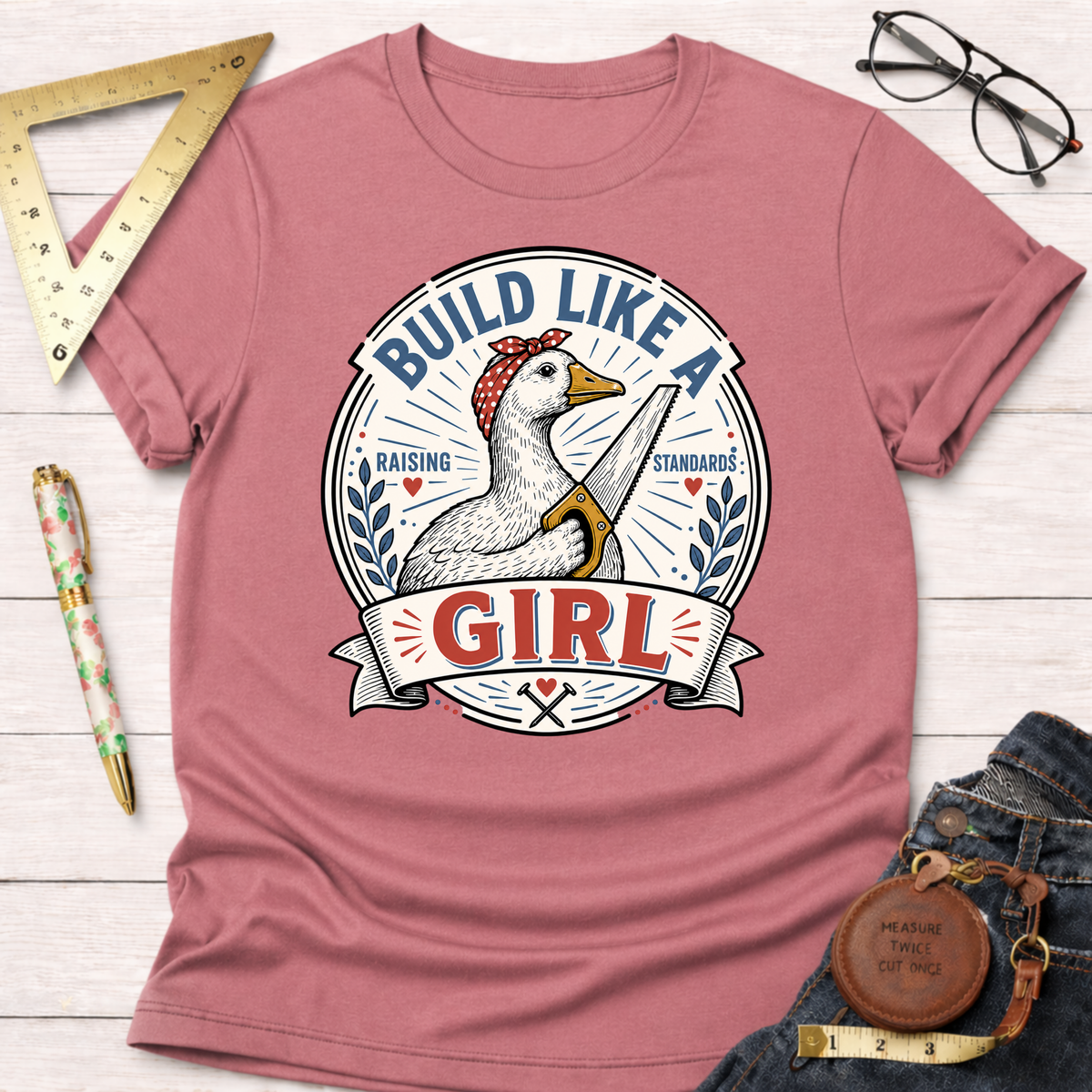 Dusty rose "Build Like A Girl" t-shirt with a goose holding a saw, surrounded by woodworking tools.