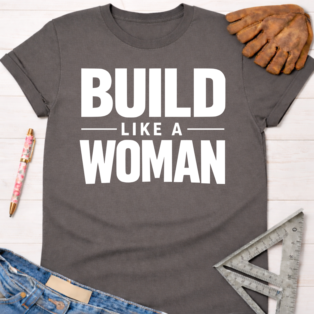 Charcoal gray "Build Like A Woman" graphic tee with white text, denim jeans, and work tools. Women's funny woodworking graphic tee shirt for female woodworkers, makers, and DIY builders — Sawdust & Ponytails.