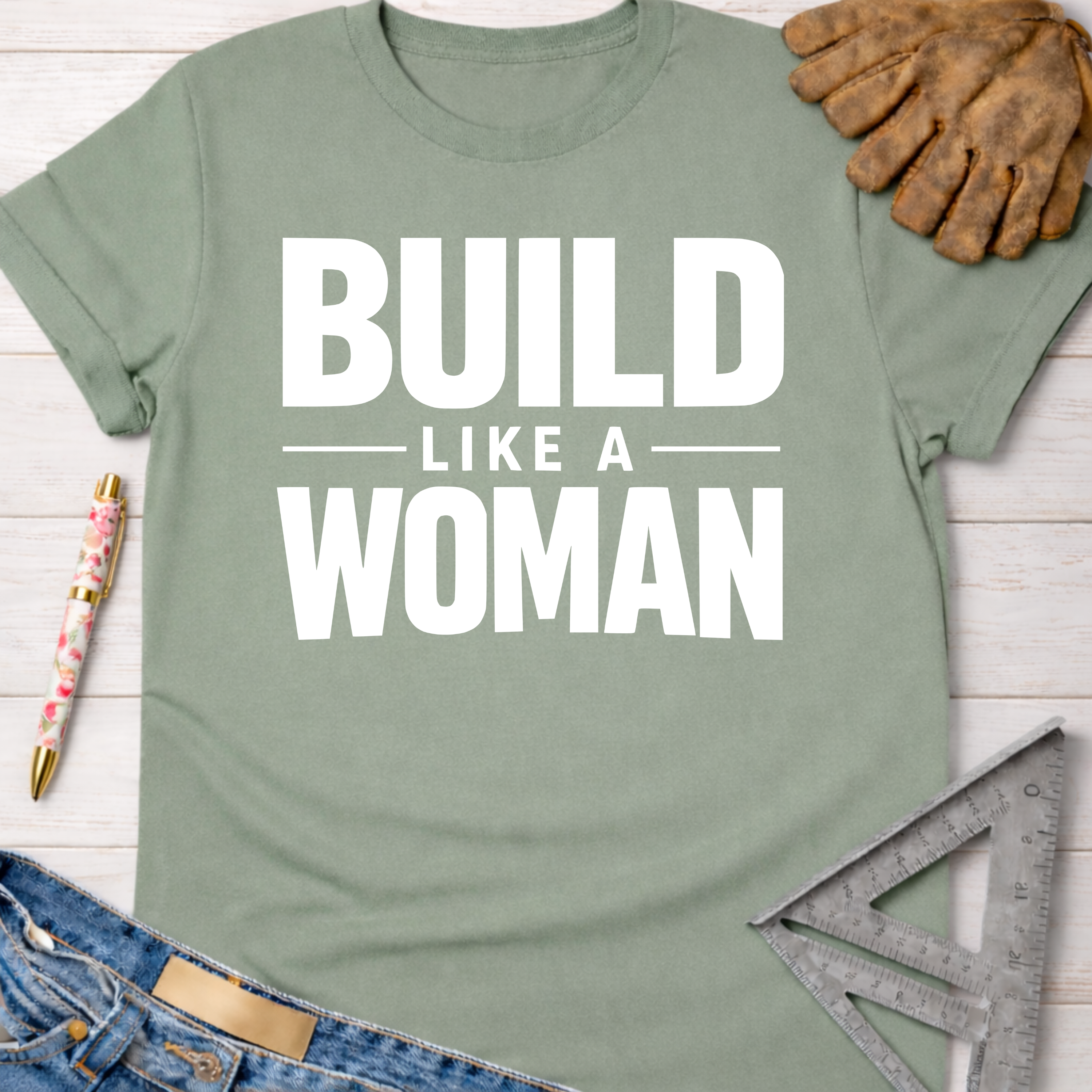 Women's funny woodworking graphic tee shirt for female woodworkers, makers, and DIY builders — Sawdust & Ponytails. Sage green cotton t-shirt with "BUILD LIKE A WOMAN" in white text.
