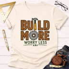 Women's funny woodworking graphic tee shirt for female woodworkers, makers, and DIY builders — Sawdust & Ponytails. Cream t-shirt with "BUILD MORE WORRY LESS" wood-textured text, hammer, and saw blade graphic.