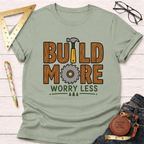 Women's funny woodworking graphic tee shirt for female woodworkers, makers, and DIY builders — Sawdust & Ponytails. Light green tee with "BUILD MORE WORRY LESS" wood-grain text, hammer, and saw blade graphic.