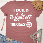 Women's funny woodworking graphic tee shirt for female woodworkers, makers, and DIY builders — Sawdust & Ponytails. Mauve t-shirt with "I Build to Fight Off The Crazy" text and crazy face emoji.