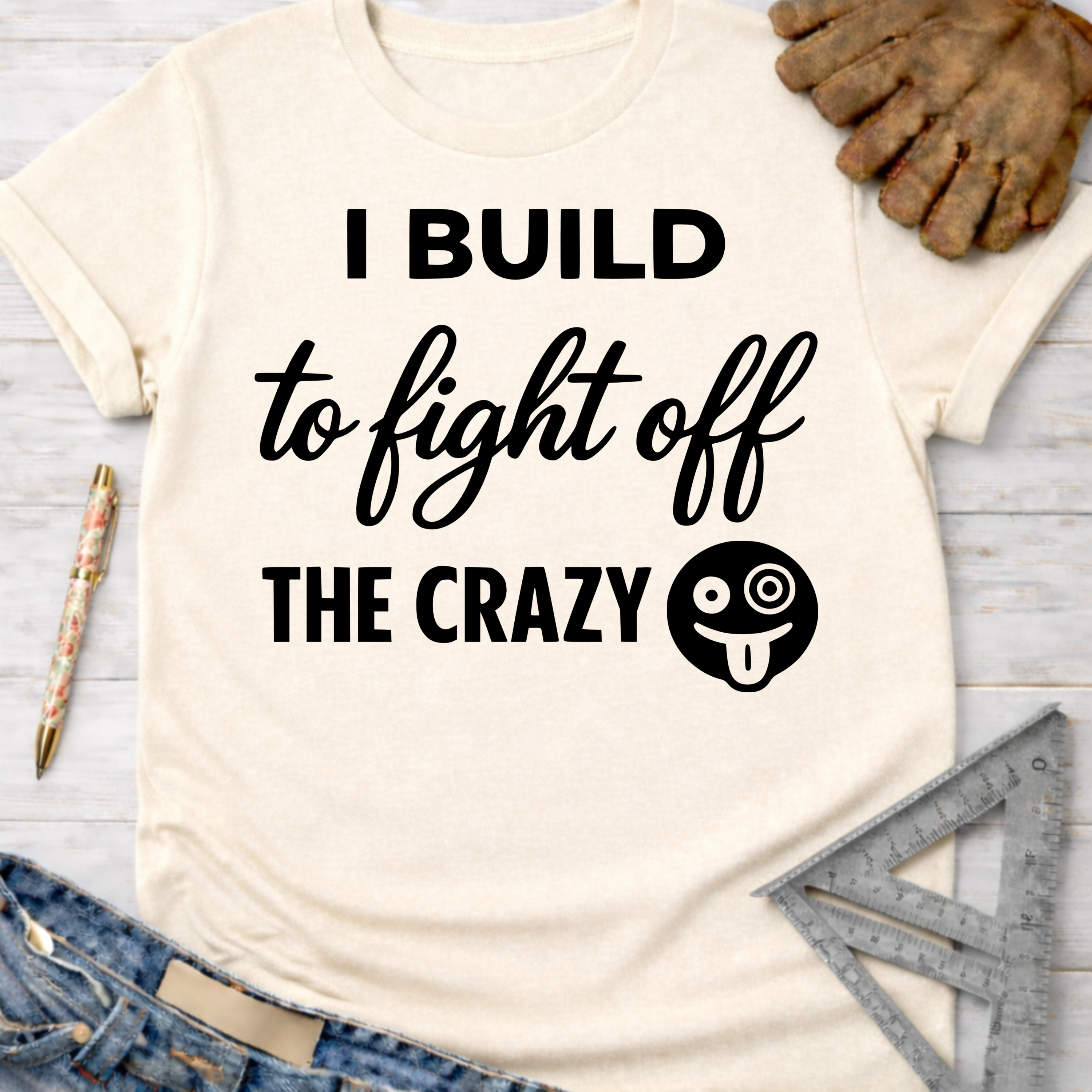 Women's funny woodworking graphic tee shirt for female woodworkers, makers, and DIY builders — Sawdust & Ponytails. Cream "I Build to Fight Off The Crazy" t-shirt with a goofy emoji.