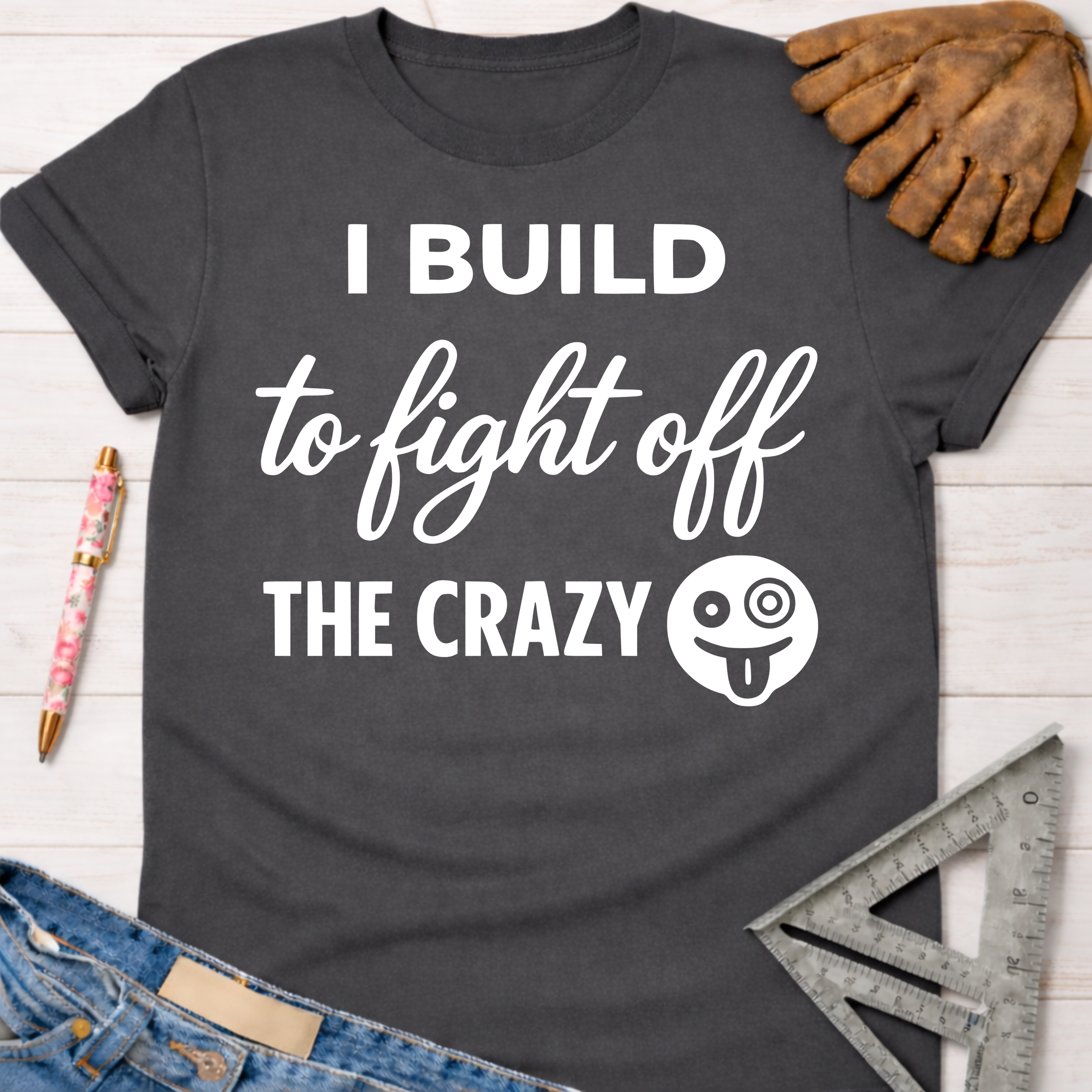 Women's funny woodworking graphic tee shirt for female woodworkers, makers, and DIY builders — Sawdust & Ponytails. Dark gray cotton tee with "I BUILD to fight off THE CRAZY" text and a silly face graphic.