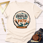 Cream t-shirt with "Build Your Own Path" design featuring a tree ring, saw, plane, and mountains.