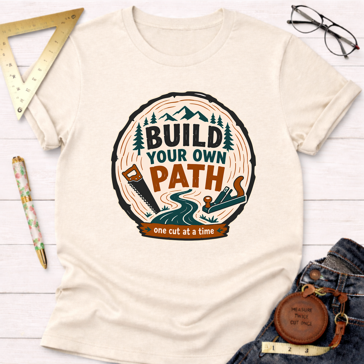 Cream t-shirt with "Build Your Own Path" design featuring a tree ring, saw, plane, and mountains.