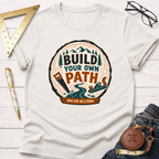 Cream t-shirt with "Build Your Own Path" wood slice graphic, featuring trees, saw, plane, and a winding road.