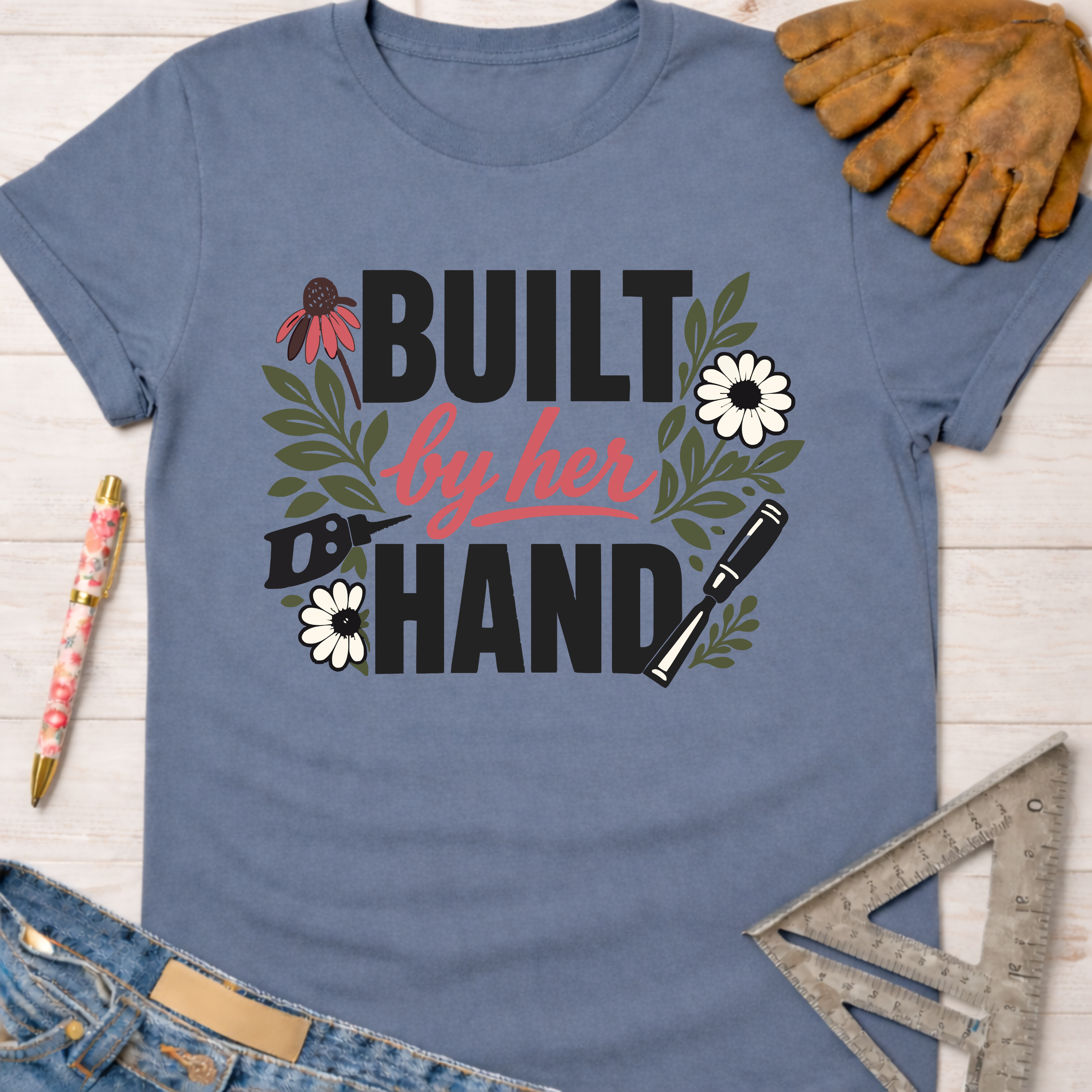 Women's funny woodworking graphic tee shirt for female woodworkers, makers, and DIY builders — Sawdust & Ponytails. Blue t-shirt with "Built By Her Hand" text, flowers, saw, and chisel.