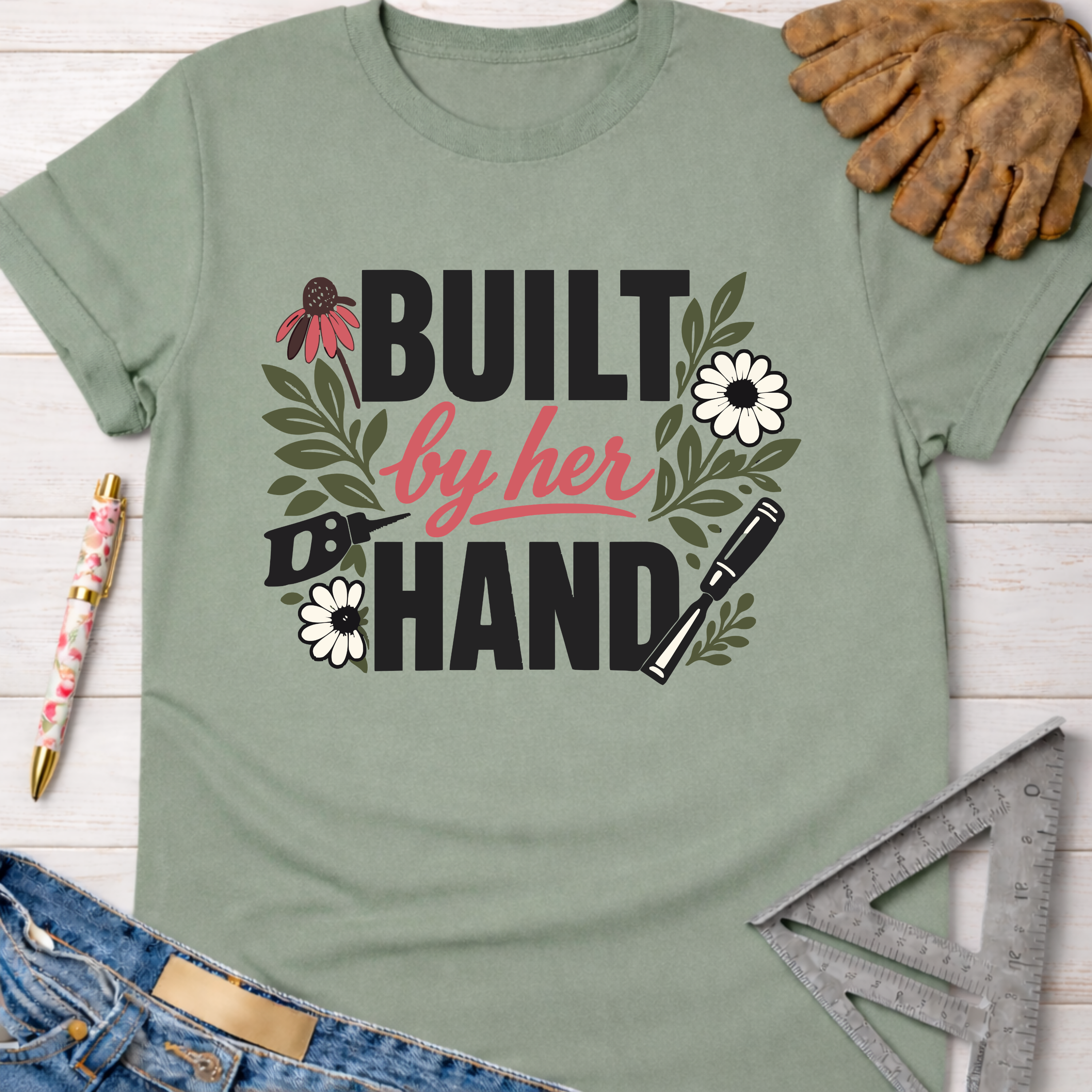 Women's funny woodworking graphic tee shirt for female woodworkers, makers, and DIY builders — Sawdust & Ponytails. Sage green cotton t-shirt with "Built by Her Hand" graphic, floral and tool designs.