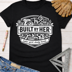 Black cotton "Built By Her" graphic tee with tools, flowers, and "maker spirit, steady hands" text. Women's funny woodworking graphic tee shirt for female woodworkers, makers, and DIY builders — Sawdust & Ponytails.