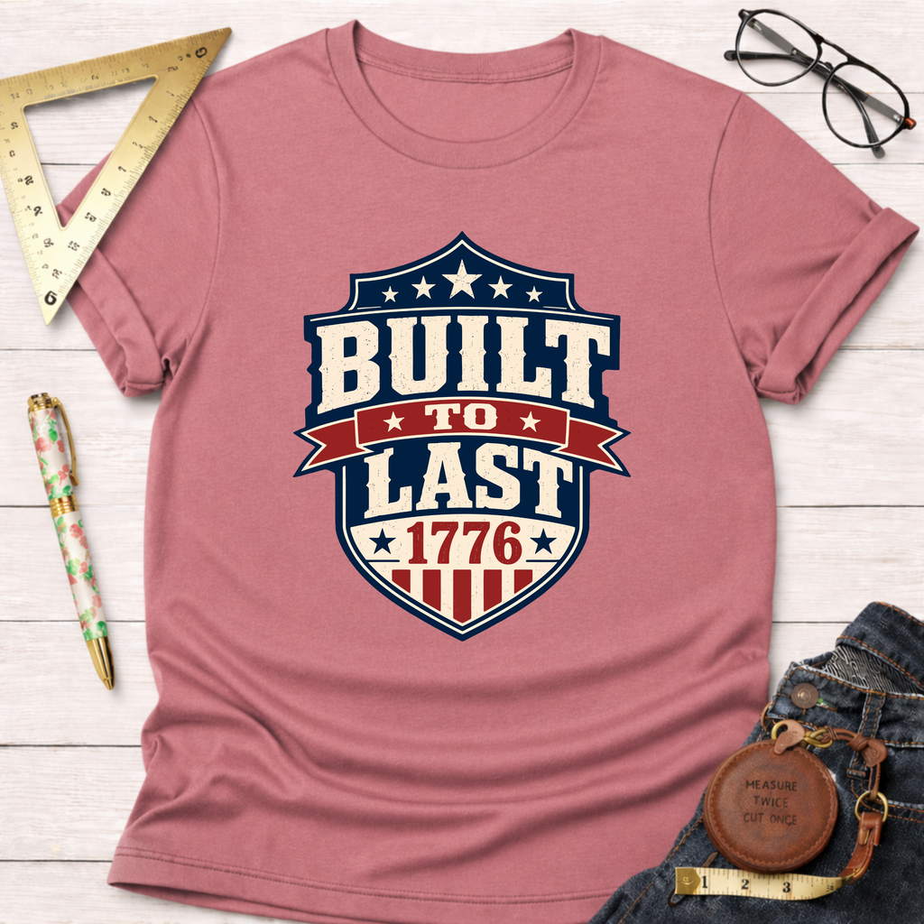 Pink "Built To Last 1776" badge t-shirt with patriotic design, styled with jeans, ruler, and glasses.