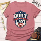 Pink "Built To Last 1776" badge t-shirt with patriotic design, styled with jeans, ruler, and glasses.