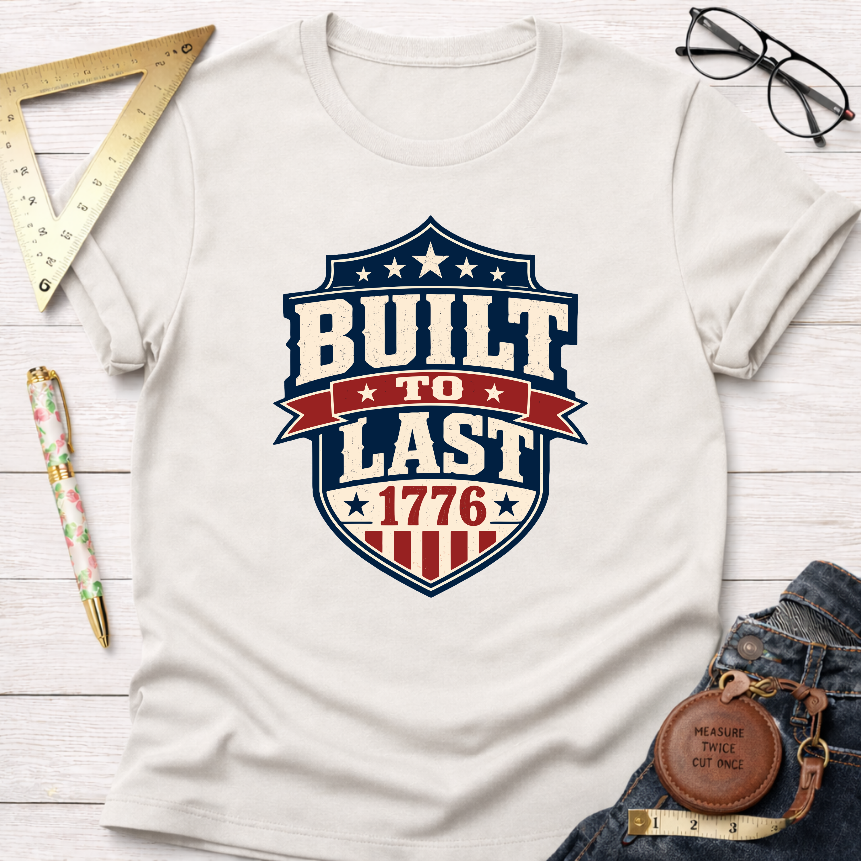 Cream cotton t-shirt with a "Built To Last 1776" badge design in red, white, and blue.