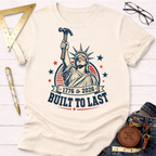 Cream t-shirt with a Lady Liberty graphic holding a hammer, "1776 to 2026 Built To Last" text.