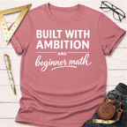 Women's funny woodworking graphic tee shirt for female woodworkers, makers, and DIY builders — Sawdust & Ponytails. Mauve t-shirt with "Built With Ambition And Beginner Math" in white text.